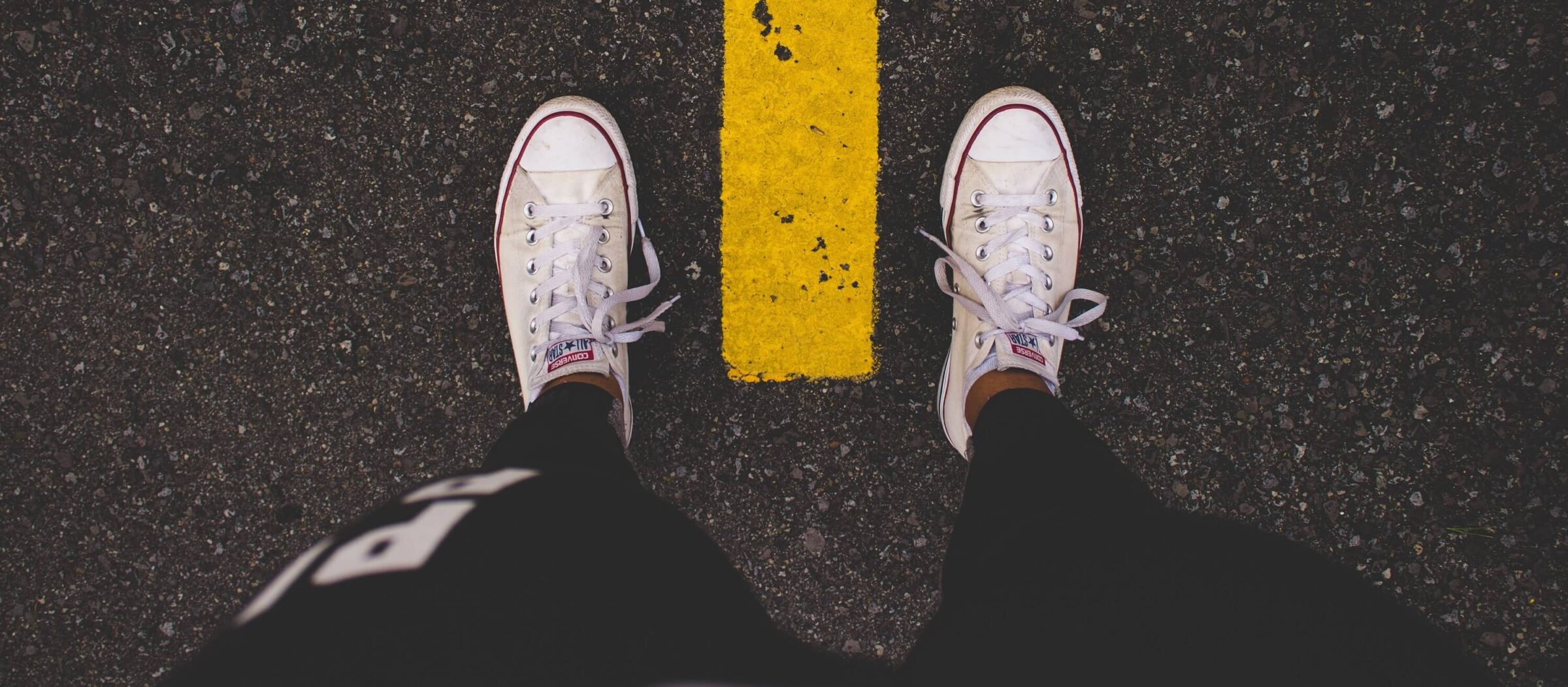 tops of white sneakers straddling yellow line on black pavement